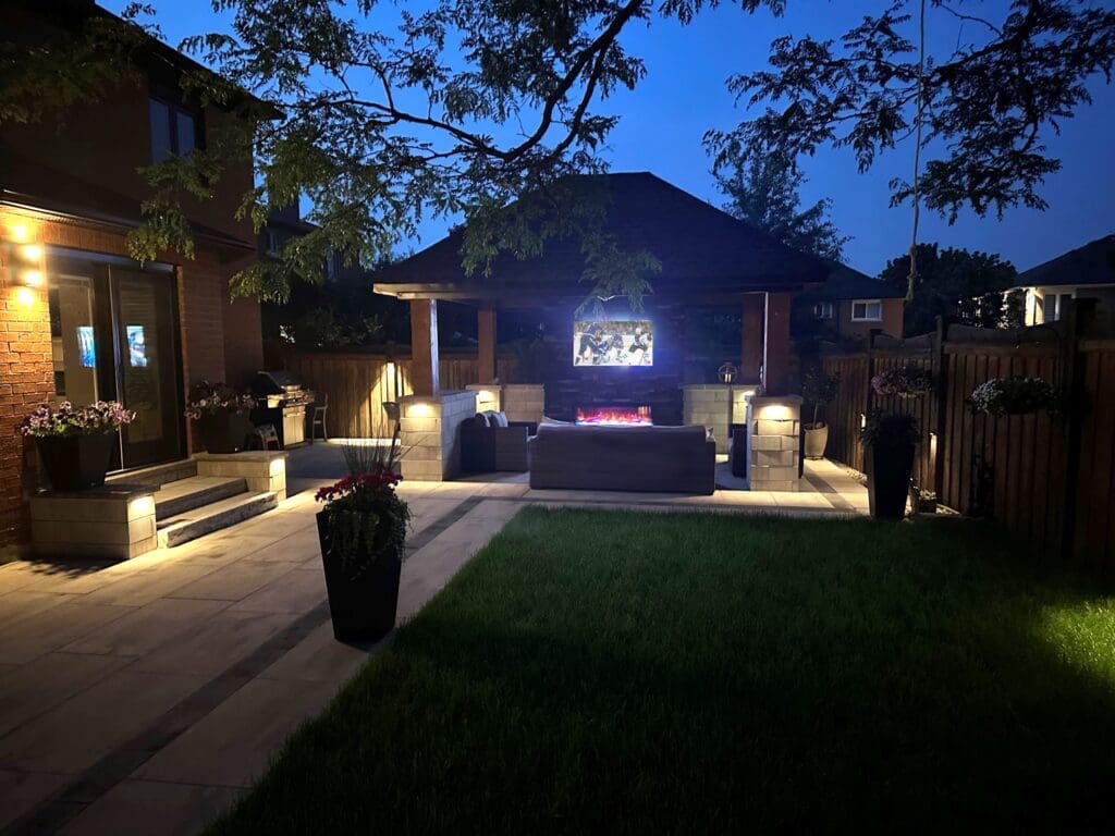 Backyard gazebo with fireplace, TV and outdoor lounge lit at night